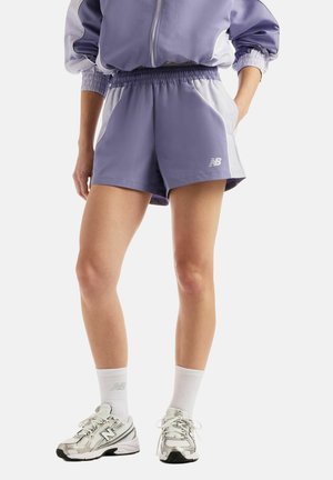 Kvinde iført lilla atletikshorts, matchende jakke, hvide sokker og sølvfarvede New Balance løbesko, står med den ene hånd i lommen.