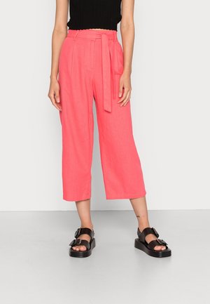 Kobieta nosząca koralowo-różowe, szerokie spodnie z wysokim stanem, sięgające do połowy łydki, z pasującym materiałowym paskiem oraz czarne sandały na platformie z klamrami.