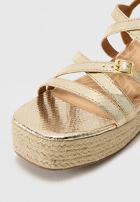 Sandale plateforme dorée métallique avec des brides entrecroisées, un dessus texturé et une semelle en jute tressé. Elle possède une boucle pour un maintien sécurisé.