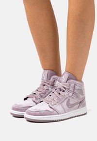 Světlé fialové semišové vysoké tenisky s saténovými tkaničkami, mají strukturovanou část swoosh a logo, a bílou gumovou podrážku.