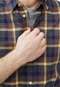 Camicia a quadri abbottonata in blu scuro, giallo e marrone. Mano del modello che stringe il colletto. Include una maglietta grigia sotto.