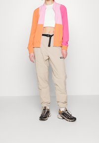 Veste colorée en orange, rose et rose clair avec un design court ; pantalon beige avec une ceinture noire et un détail logo ; baskets noires et fauves.