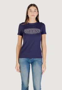 Maglietta in cotone blu navy con logo "GUESS" in strass sul fronte. Maniche corte e vestibilità classica. Indossata con jeans azzurri chiari.