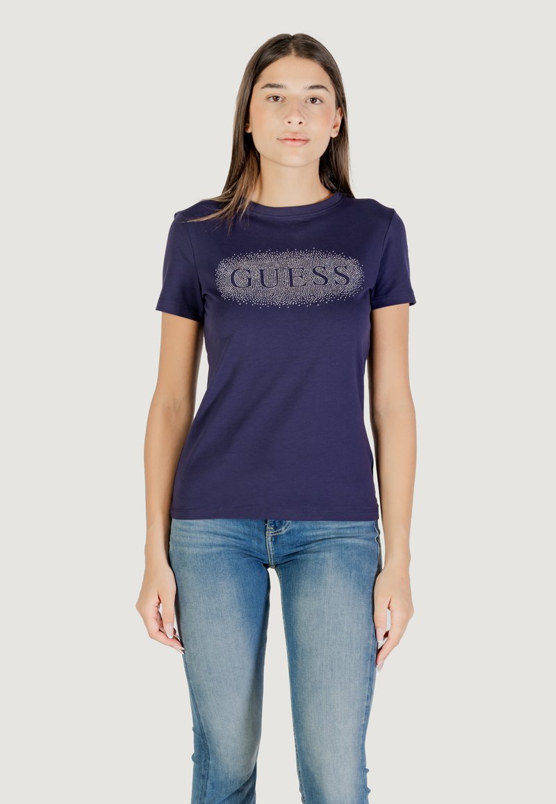 Maglietta in cotone blu navy con logo "GUESS" in strass sul fronte. Maniche corte e vestibilità classica. Indossata con jeans azzurri chiari.