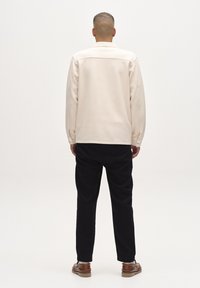 Pikad käised, heledast beežist särgist, millel on tekstuuriga viimistlus ja nööpidega mansetid, koos mustade pükste ja pruunide kingadega, vaadatuna tagant.