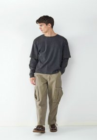 Donkergrijze oversized longsleeve shirt gecombineerd met lichtkhaki cargobroek met zijzakken. Bruine veterschoenen maken de outfit compleet.