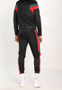 Svart träningsdräkt med röda och gröna detaljer, med en figurnära design och elastiska muddar. Tillverkad av ett slätt, glänsande material. Vita sneakers.