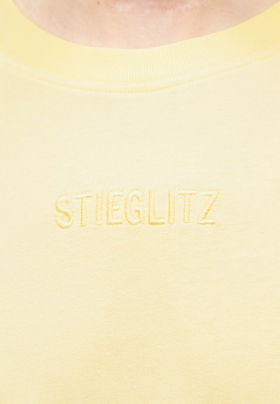 Κοντινό πλάνο ενός κίτρινου πουλόβερ με στρογγυλό λαιμόκοψη και ανάγλυφο κεντητό κείμενο "STIEGLITZ" στο στήθος.