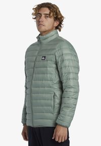 Leichte grüne gepolsterte Jacke mit vertikaler Steppstruktur, hohem Kragen, Seitentaschen und kleinem Logo auf der linken Brust.