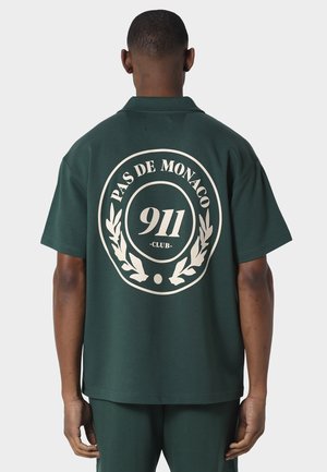 Mann trägt ein dunkelgrünes Kurzarmhemd mit großem weißem "Pas de Monaco 911 Club"-Logo und Lorbeerkranz auf dem Rücken.
