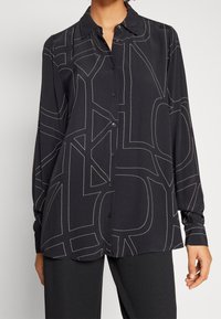 Maglietta nera a maniche lunghe con cuciture geometriche bianche, frontale con bottoni e colletto. Tessuto liscio con vestibilità comoda.