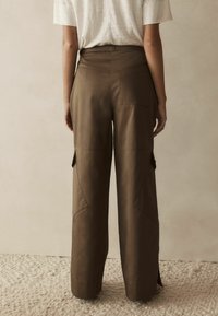 Pantaloni cargo maro, cu croi larg, dotati cu buzunare frontale, cusături vizibile și o textură netedă. Viziune din spate arată un design cu talie înaltă.