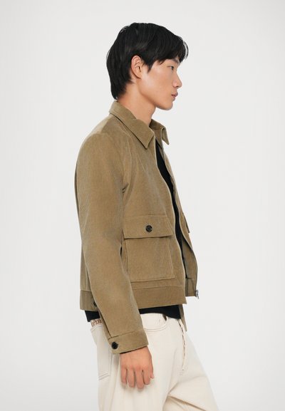 Brun corduroyjakke med krave, forlommer og knaplukning, båret over en sort skjorte og parret med cremede bukser.