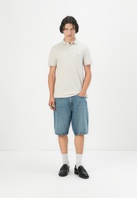 Cămașă polo bej cu mâneci scurte și logo discret, asociată cu pantaloni scurți din denim albastru, șosete gri și mocasini negri. Outfit simplu, casual.