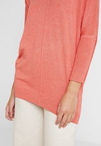 Korali sweter dzianinowy z prążkowanymi mankietami, opuszczonymi ramionami i bokiem z rozcięciem. Połączony z jasnobeżowymi spodniami o szerokich nogawkach.