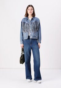 Jeansjacke mit Fransen in Hellblau, kombiniert mit weiten blauen Jeans und weißen Sneakers, hält eine kleine olivgrüne Tasche.