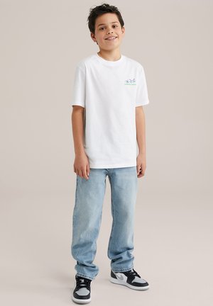 T-shirt bianca in cotone con una stampa grafica verde, abbinata a jeans in denim blu chiaro e sneakers nere con accenti grigi.