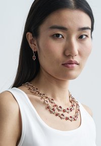 Gouden sieradenset met een gelaagde ketting voorzien van rode en heldere kristallen, vergezeld van bijpassende hangende oorbellen. Zachte en glanzende afwerking.