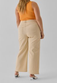 Pantalones cargo anchos beige con dos bolsillos traseros y bolsillos laterales, combinados con una camiseta sin mangas naranja y sandalias de tacón plateadas.