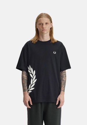 Jonge man met schouderlang blond haar, die een zwart oversized T-shirt draagt met een witte lauwerkransprint en getatoeëerde armen op een witte achtergrond.