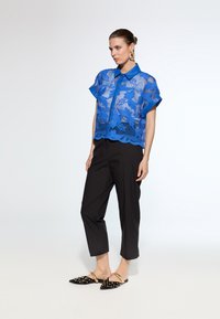 Blue lace button-up shirt with short sleeves, featuring a floral pattern and scalloped hem, paired with black tapered pants and studded flats.