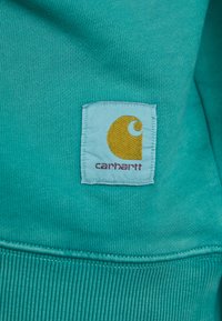 Turkise stof med ribbet tekstur og en rektangulær blå lap med et gult logo og teksten "carhartt."