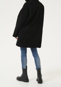 Person som bär en lång svart fleecekappa, blå slim fit-jeans och svarta läderankelboots som står framför en vit bakgrund.