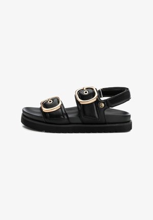 Sandalia negra con suela gruesa, que presenta dos tiras anchas con hebillas doradas. Material de cuero suave y parte trasera abierta para facilitar el calzado.