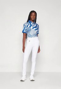 Γυναίκα με μακριές πλεξούδες, φορώντας ένα μπλε και λευκό πουκάμισο πόλο κοντομάνικο, λευκά εφαρμοστά παντελόνια και λευκά αθλητικά παπούτσια, που στέκεται σε λευκό φόντο.