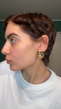 Pendientes de metal dorado en forma de discos abstractos, que reflejan la luz con una superficie lisa y bordes redondeados. Llevados con un peinado corto.