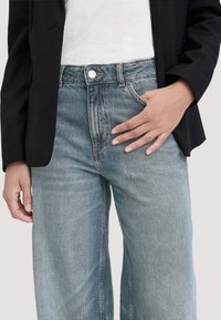 Jeans azzurri chiari in denim, con vita alta, gamba larga e design classico a cinque tasche. Un blazer nero è indossato sopra una maglietta bianca.