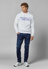 Sudadera blanca con "RED BULL RACING" impreso en púrpura. Combinada con jeans azul oscuro y zapatillas grises con detalles blancos. Estilo casual.