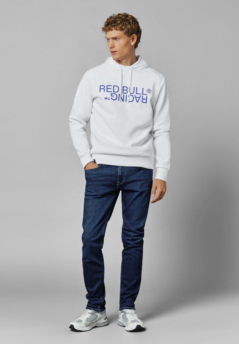 Sudadera blanca con "RED BULL RACING" impreso en púrpura. Combinada con jeans azul oscuro y zapatillas grises con detalles blancos. Estilo casual.