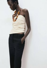 Haut beige sans bretelles avec des détails froncés, associé à un pantalon noir ajusté. Les accessoires comprennent un collier en perles multicolores.