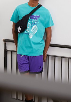 Osoba ubrana w turkusowy t-shirt z nadrukiem, fioletowe krótkie spodenki do koszykówki, czarną torebkę Nike noszoną przez ramię, stoi przy czarnej metalowej balustradzie w pomieszczeniu.