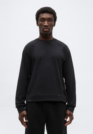 Man stands facing forward wearing a plain black long-sleeve sweatshirt and black pants against a light gray background.