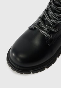 Črne usnjene škornje z debelim gumijastim podplatom in črno-bež vzorčenimi vezalkami, prikazane z zgornje bližnje perspektive.