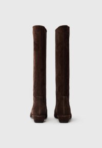 Une paire de bottes en daim brun foncé, hautes, avec de petits talons carrés, vues de l'arrière sur un fond léger et uni.