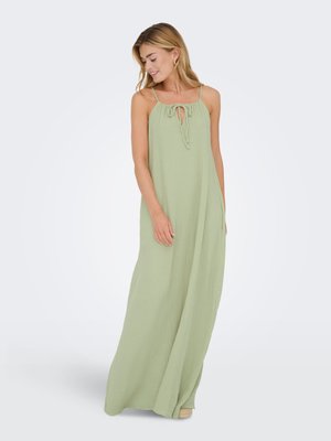 Donna che indossa un abito lungo verde chiaro senza maniche con spalline sottili e scollo a goccia annodato, in piedi e che guarda verso il basso.