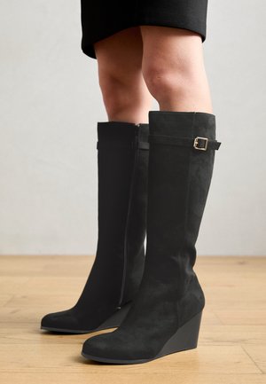 Kobieta w czarnych zamszowych kozaczkach na koturnie sięgających do kolan, z bocznym zamkiem i złotym paskiem z klamrą, stojąca na drewnianej podłodze.