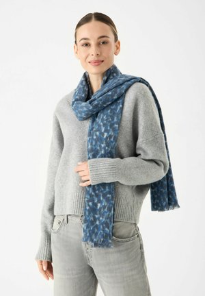 Écharpe à motif bleu en tissu doux, drapée autour du cou, associée à un pull léger gris et un jean gris. Les extrémités à franges sont visibles.
