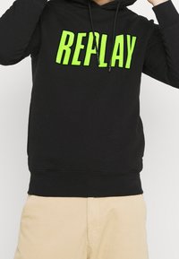 Sudadera negra con un gran estampado en verde brillante de "REPLAY". Cuenta con capucha con cordón y dobladillo de canalé, fabricada en una tela suave.