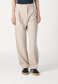 Beige wijde broek met voorplooien, gemaakt van een soepele stof. Gecombineerd met zwarte sneakers met een rubberen zool en paarse accenten.