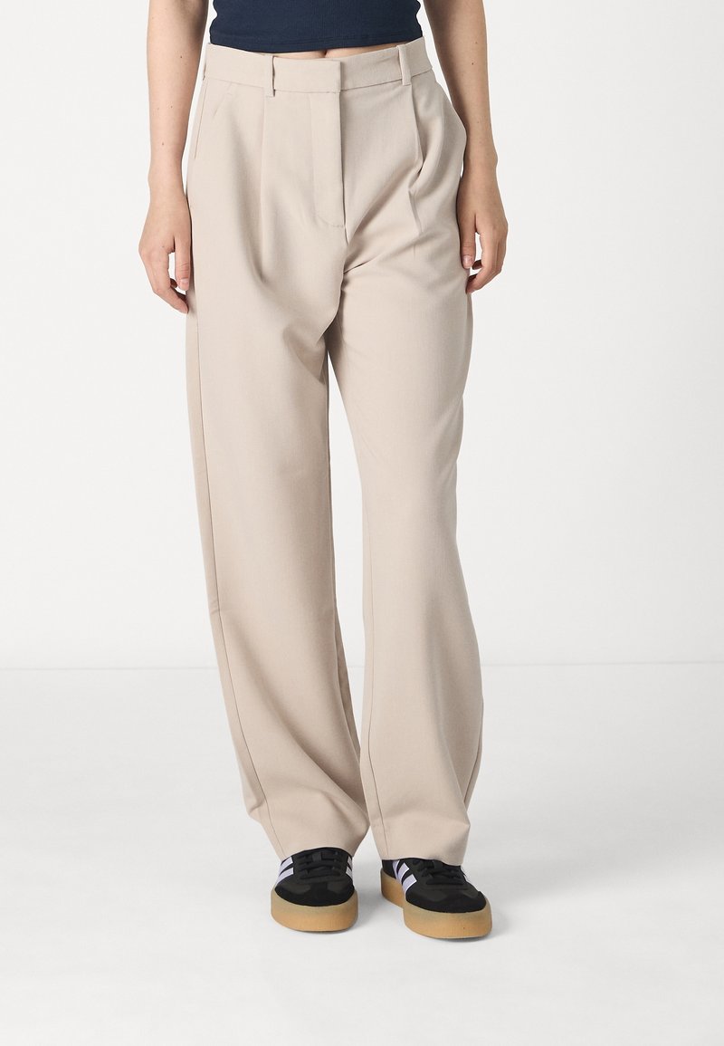 Beige wijde broek met voorplooien, gemaakt van een soepele stof. Gecombineerd met zwarte sneakers met een rubberen zool en paarse accenten.