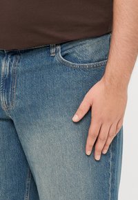 Heledad hall värvi denim Jeanid, kerge kulumisefektiga, esiküljel tasku ja pruuni värvi vöökoht, millel on nööp. Käsi toetub reitele.