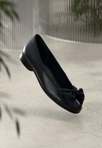 Chaussure plate en cuir noir avec un bout rond et un grand nœud décoratif, flottant au-dessus d'un sol en béton clair avec un arrière-plan flou.