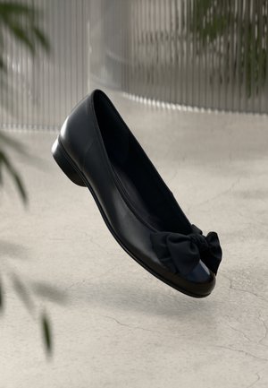 Black leather flat shoe with rounded toe and large decorative bow, floating above a light concrete floor with blurred background.