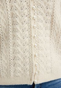 Krämfärgad stickad cardigan med kabelmönster, ribbad nederkant och pärlknappar längs framsidan. Mjuk textur.