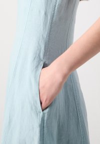 Main placée dans la poche latérale d'une robe bleu clair sans manches en tissu texturé.