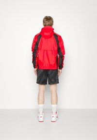 Veste légère rouge avec accents noirs, poignets élastiques et capuche, associée à un short noir. Le mannequin porte des baskets blanches et des chaussettes.